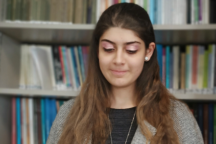 Lucia è fotografata all'interno di una biblioteca e si vedono gli scaffali pieni di libri dietro di lei. In questa foto è inquadrata a mezzo busto. È vestita in modo formale. Sta sorridendo. 