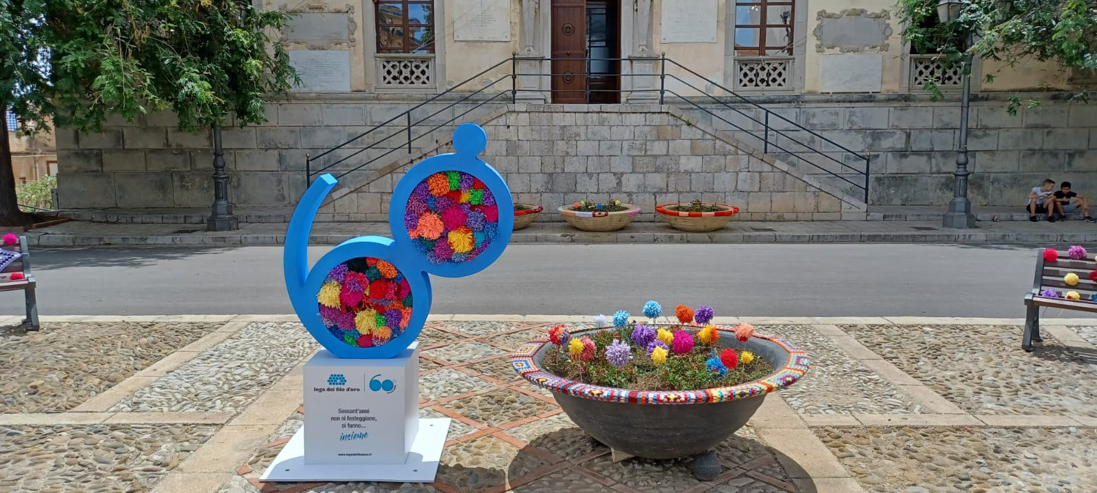 Dettaglio di un’aiuola decorata per lo Yarn Bombing: fiori colorati fatti a maglia arricchiscono il verde. Accanto, l’opera per i 60 anni della Lega del Filo d’Oro, composta da un cubo bianco con il logo e il numero 60 blu decorato con pompon multicolori.