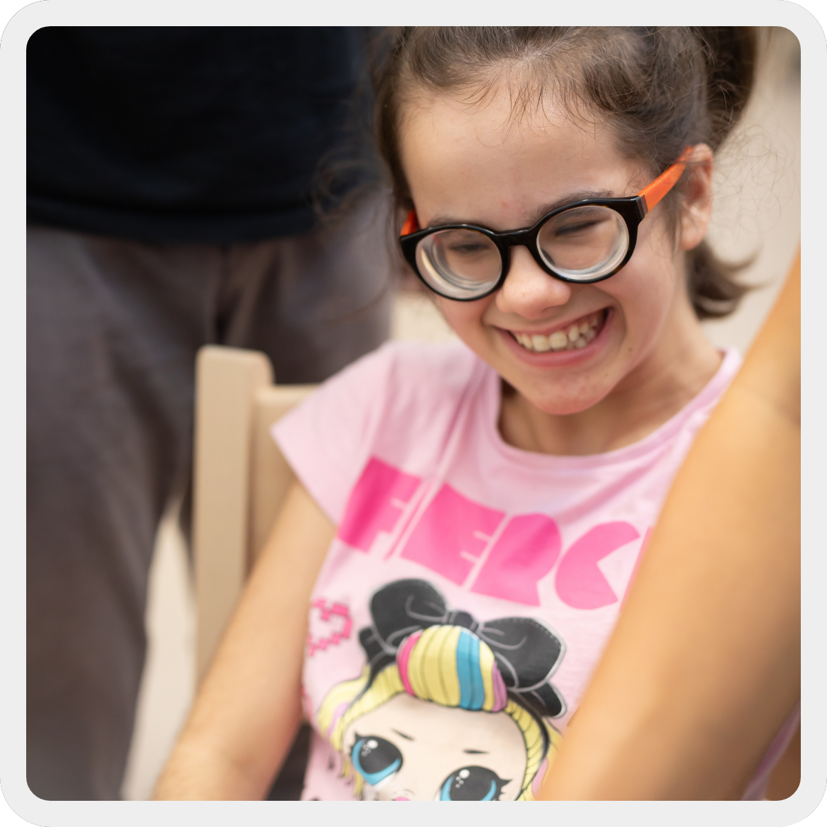 Eleonora, ragazzina con pluridisabilità di circa 15 anni, sorride divertita. Indossa una maglia rosa con un disegno di una bambola e degli occhiali tondi neri ed arancioni. 