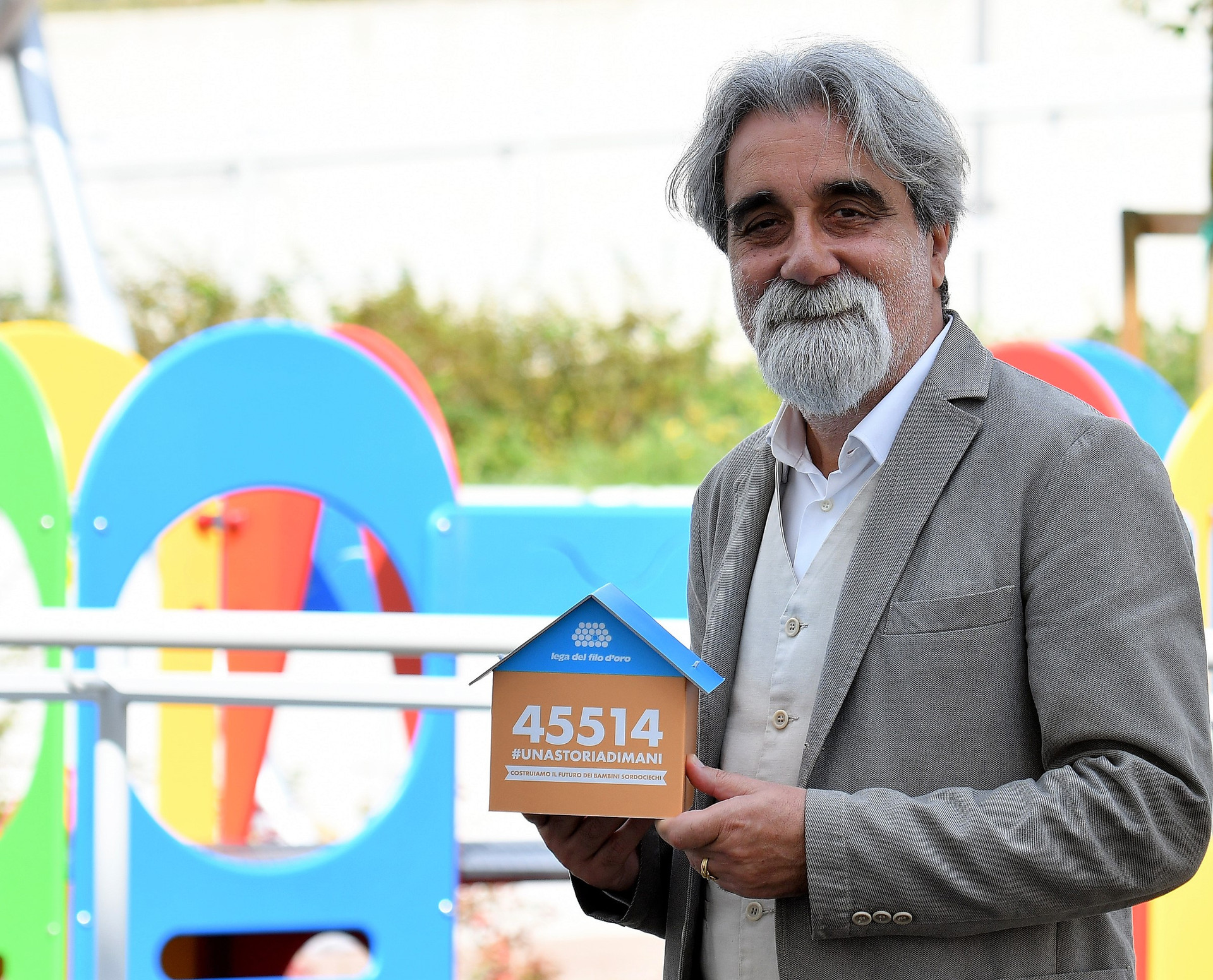 Il Maestro Beppe Vessicchio, nel parco giochi del Centro Nazionale, tiene in mano la casetta segnalino della campagna #unastoriadimani con il numero per donare via SMS solidale