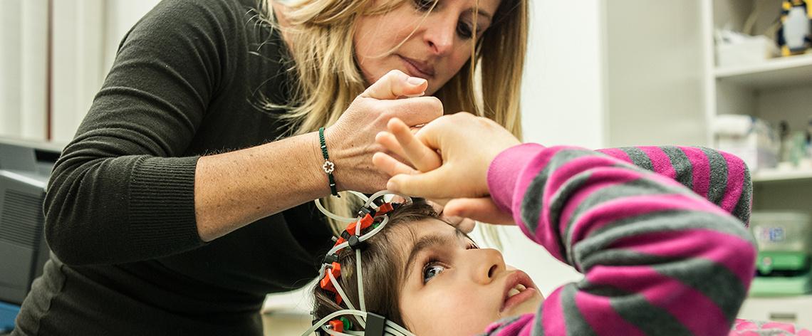 Una visita nel Laboratorio neurologico del Centro di Osimo. Nell'immagine ci sono una bambina, che indossa un maglione a righe grigie e fucsia, a cui una donna con i capelli lisci e biondi sta applicando degli elettrodi alla testa per eseguire un esame diagnostico.