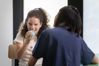 Giulia, ragazzina seguita dal Centro di Osimo, tiene in mano una pallina celeste e ride di gusto mentre svolge un esercizio seduta di fronte alla sua operatrice, che è vista di spalle nella foto.