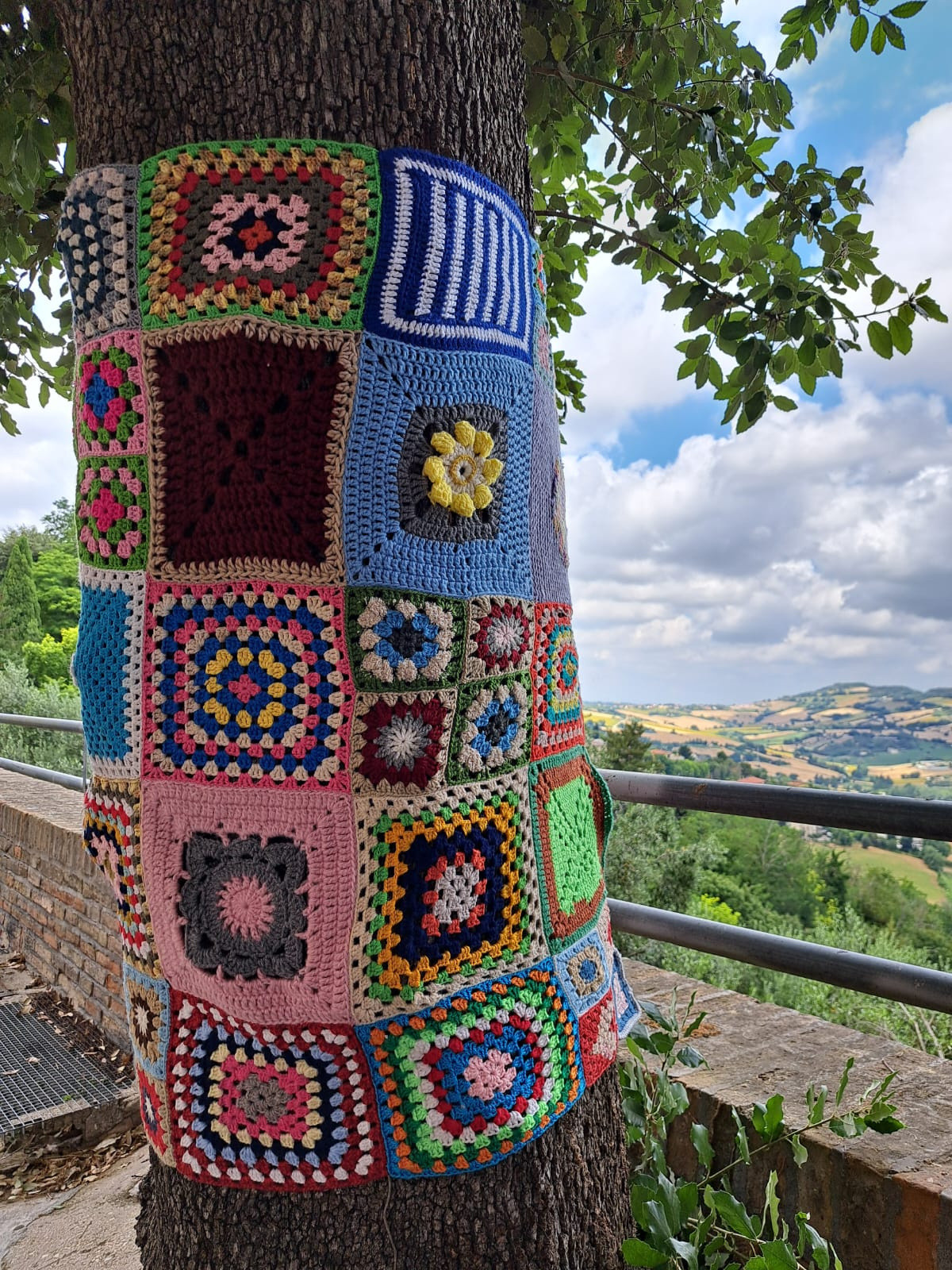 Dettaglio di un albero decorato con una grande maglia fatta di mattonelle colorate all’uncinetto, parte dello Yarn Bombing promosso dalla Lega del Filo d’Oro. Sullo sfondo si vedono colline verdi e un cielo azzurro con grandi nuvole bianche. 