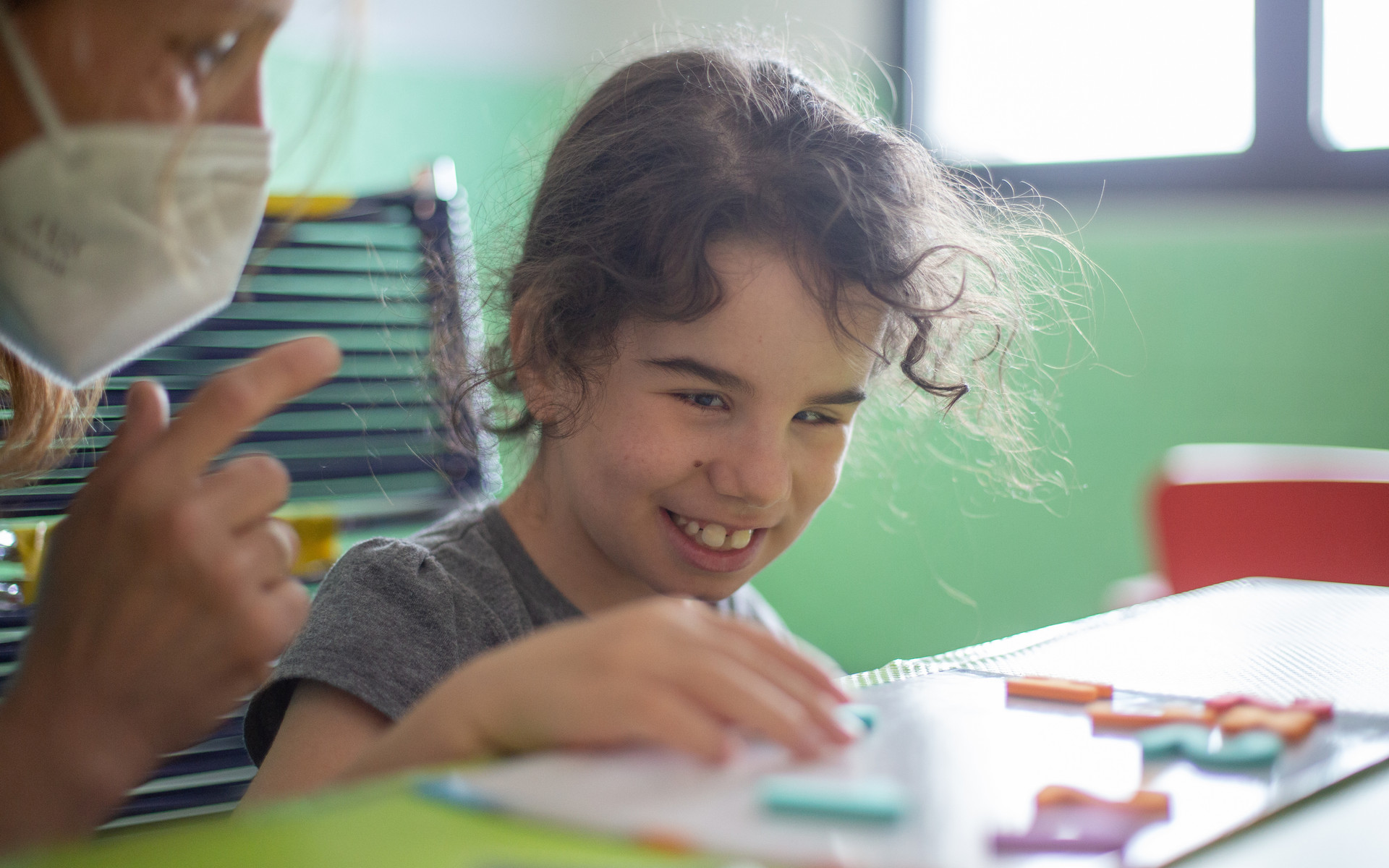 La piccola Giulia, utente della Lega, sorride mentre è al lavoro con l'educatrice davanti ad un tavolo e sposta lettere e numeri calamitati