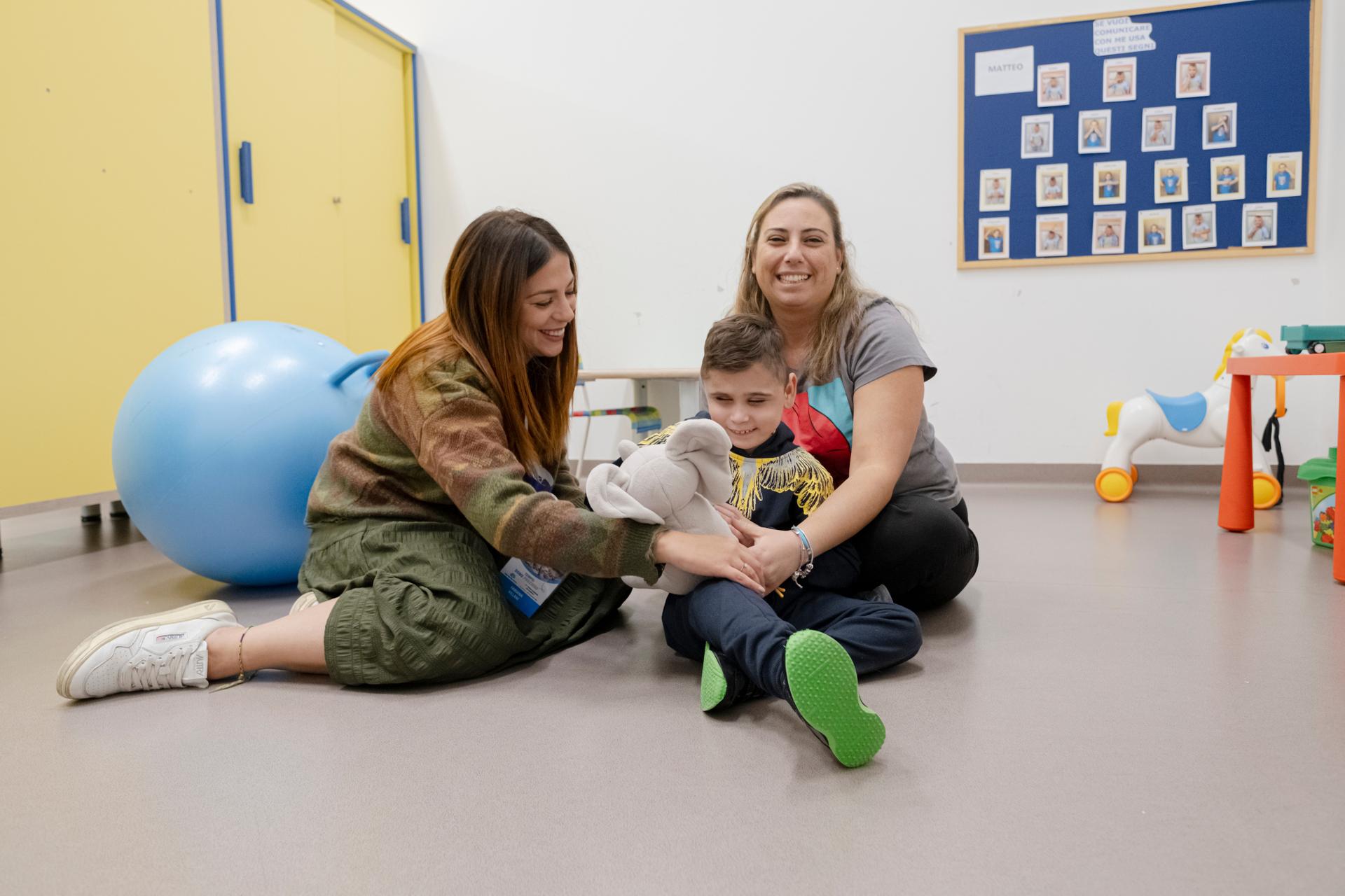 Metteo, con mamma e assistente sociale della Sede Territoriale di Napoli. Sono seduti per terra, Matteo tiene in mano un elefantino di Peluches. Sorridono