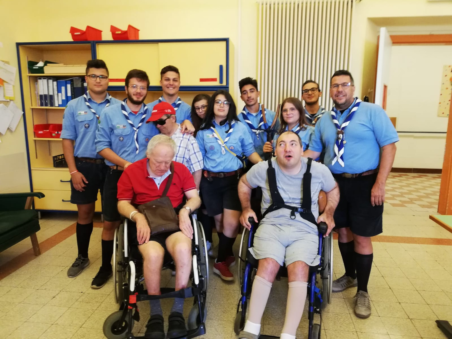 Foto di gruppo all'interno di una stanza della storica sede della Lega del Filo d'Oro di Osimo durante uno dei campi di servizio : 9 ragazzi e ragazze scout sorridono insieme a 3 utenti adulti storici, Angelo e Cristian in carrozzina e Santo 