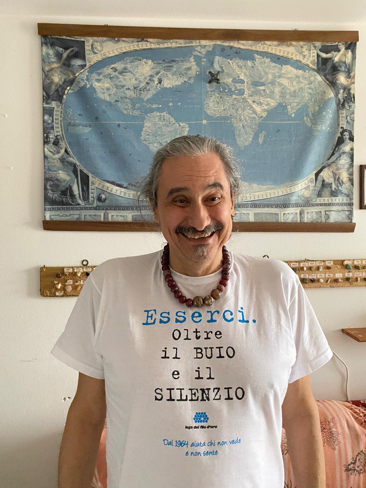 Roberto Infante con la maglia bianca della Lega del Filo d'Oro. Sulla maglia c'è la scritta "esserci oltre il buio e il silenzio". Roberto è ritratto in modo frontale e sorride felice. Sullo sfondo c'è una carta geografica appesa al muro.