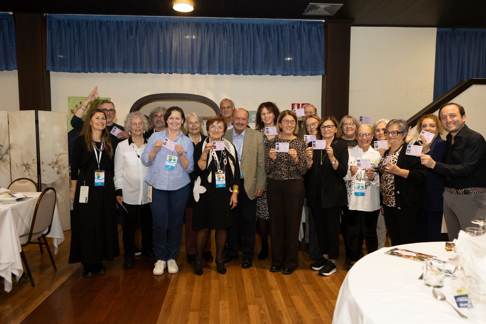 Foto di gruppo durante la cena del Forum dei Volontari. I volontari più anziani mostrano le patenti simboliche ricevute come riconoscimento per il loro lungo impegno e prezioso contributo nelle attività di volontariato.