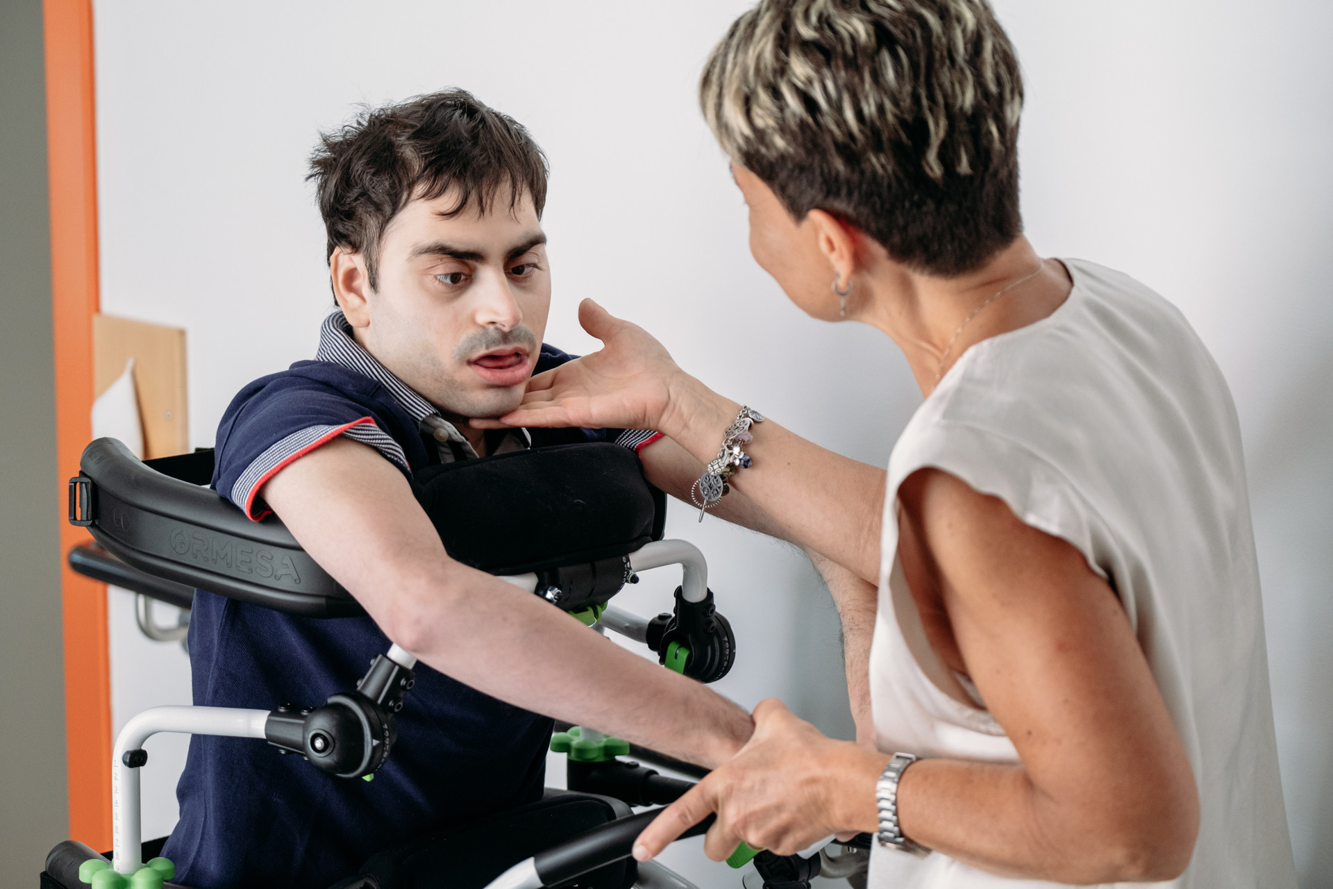 Simone, un utente adulto della Lega del Filo d'Oro è ritratto insieme alla sua educatrice mentre fa un esercizio motorio. Simone è in piedi sorretto da un deambulatore studiato appositamente per lui. 