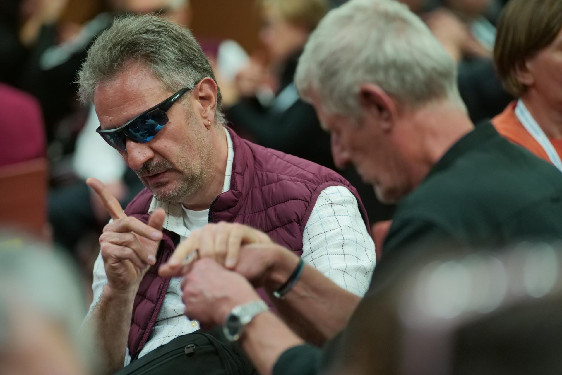 Alessandro Romano all'Assemblea delle Famiglie 2023 a Tivoli mentre parla con la LIS tattile insieme ad un'altra persona sordocieca