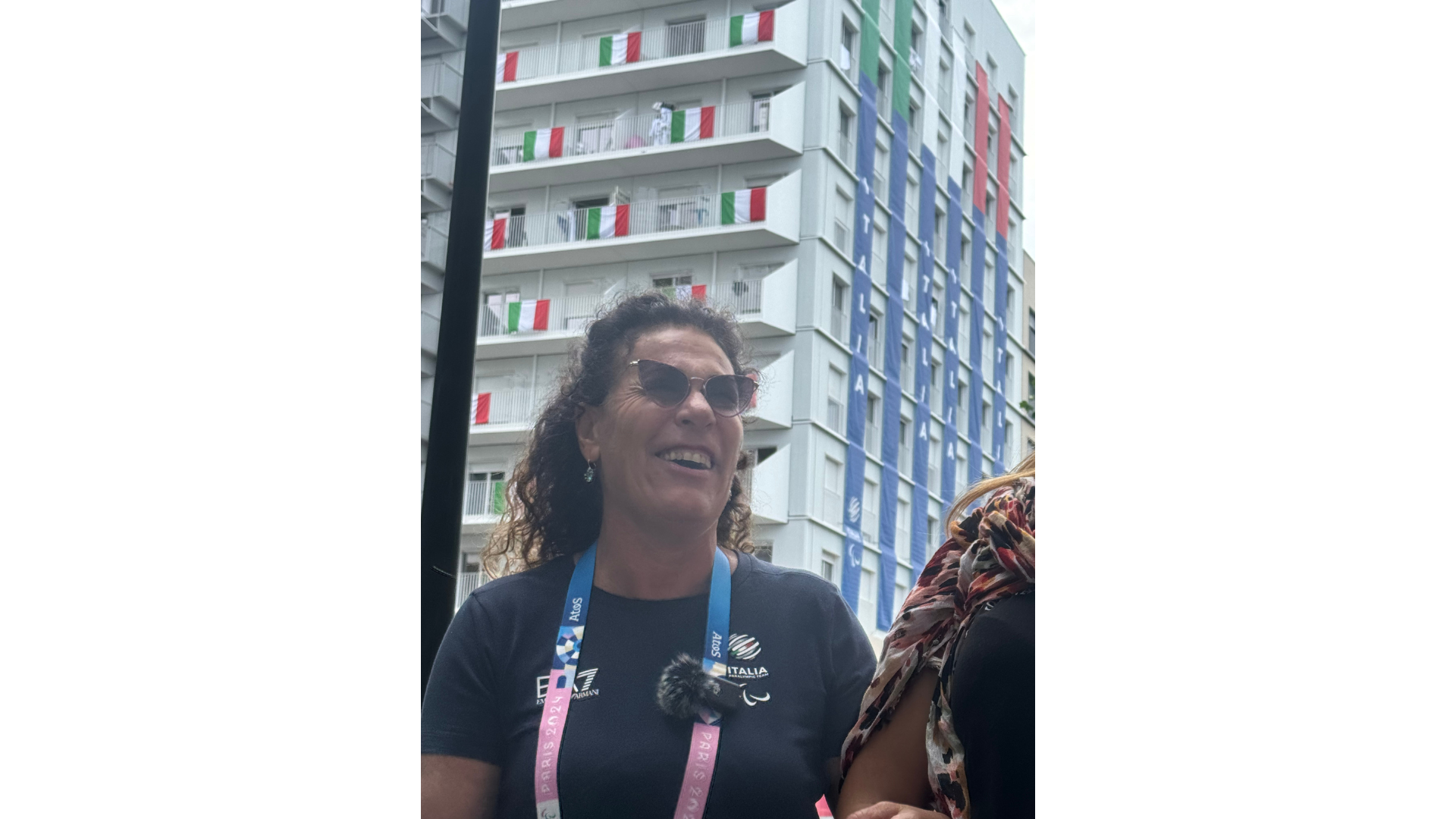 Primo piano della judoka sordocieca Matilde Lauria, sorridente durante le Paralimpiadi di Parigi 2024. Sullo sfondo si intravedono i palazzi del villaggio italiano, decorati con bandiere tricolori. 