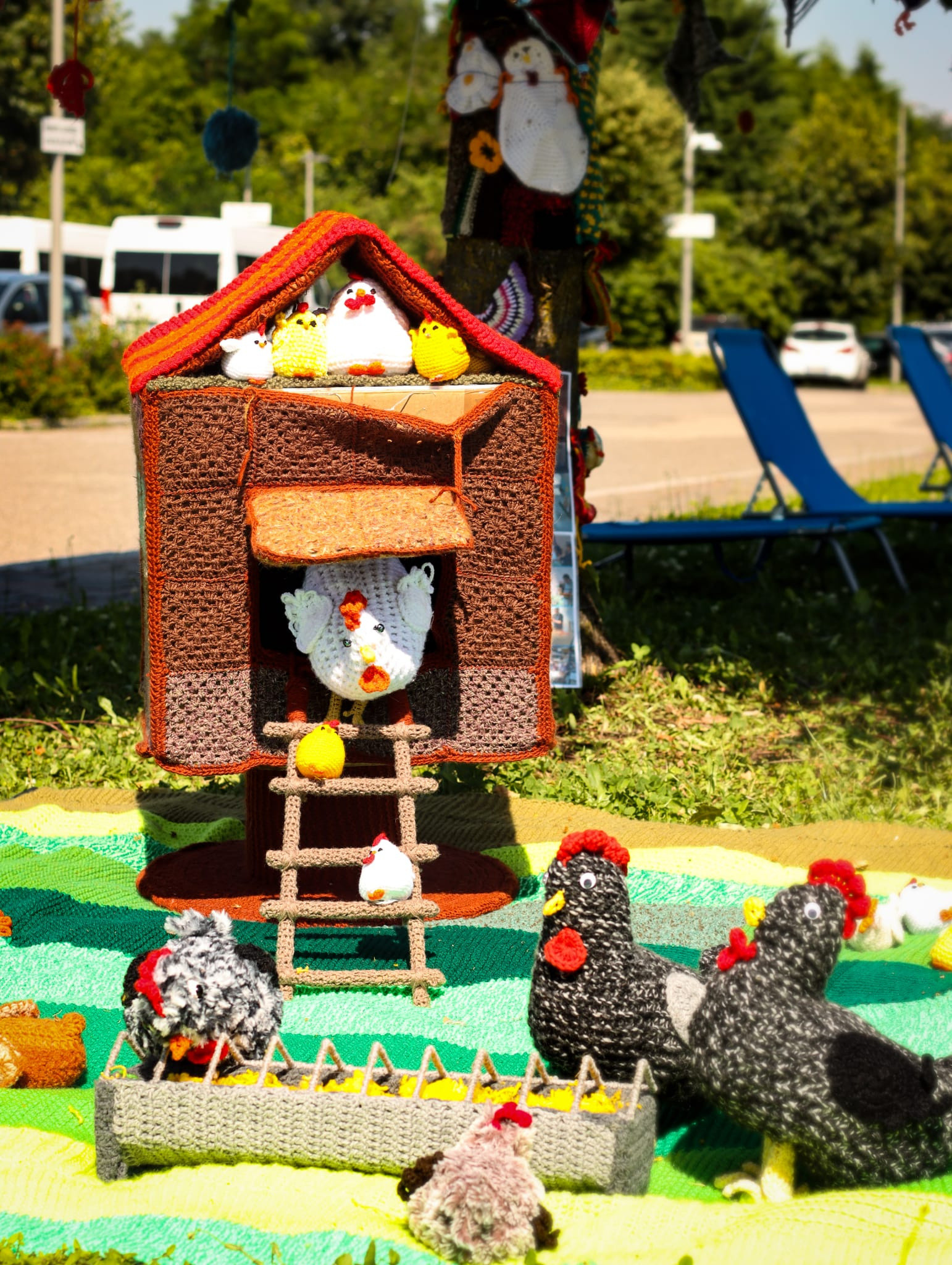 Dettaglio di un pollaio realizzato interamente all’uncinetto: presenti galli, galline, pulcini e un contenitore per il becchime, tutti fatti a mano con filati colorati. L’opera, parte dello Yarn Bombing della Lega del Filo d’Oro.