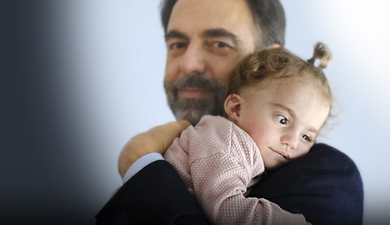Neri Marcorè tiene in braccio Benedetta, una giovane utente della Lega del Filo d’Oro. Benedetta è appoggiata alla sua spalla, avvolta in un abbraccio.