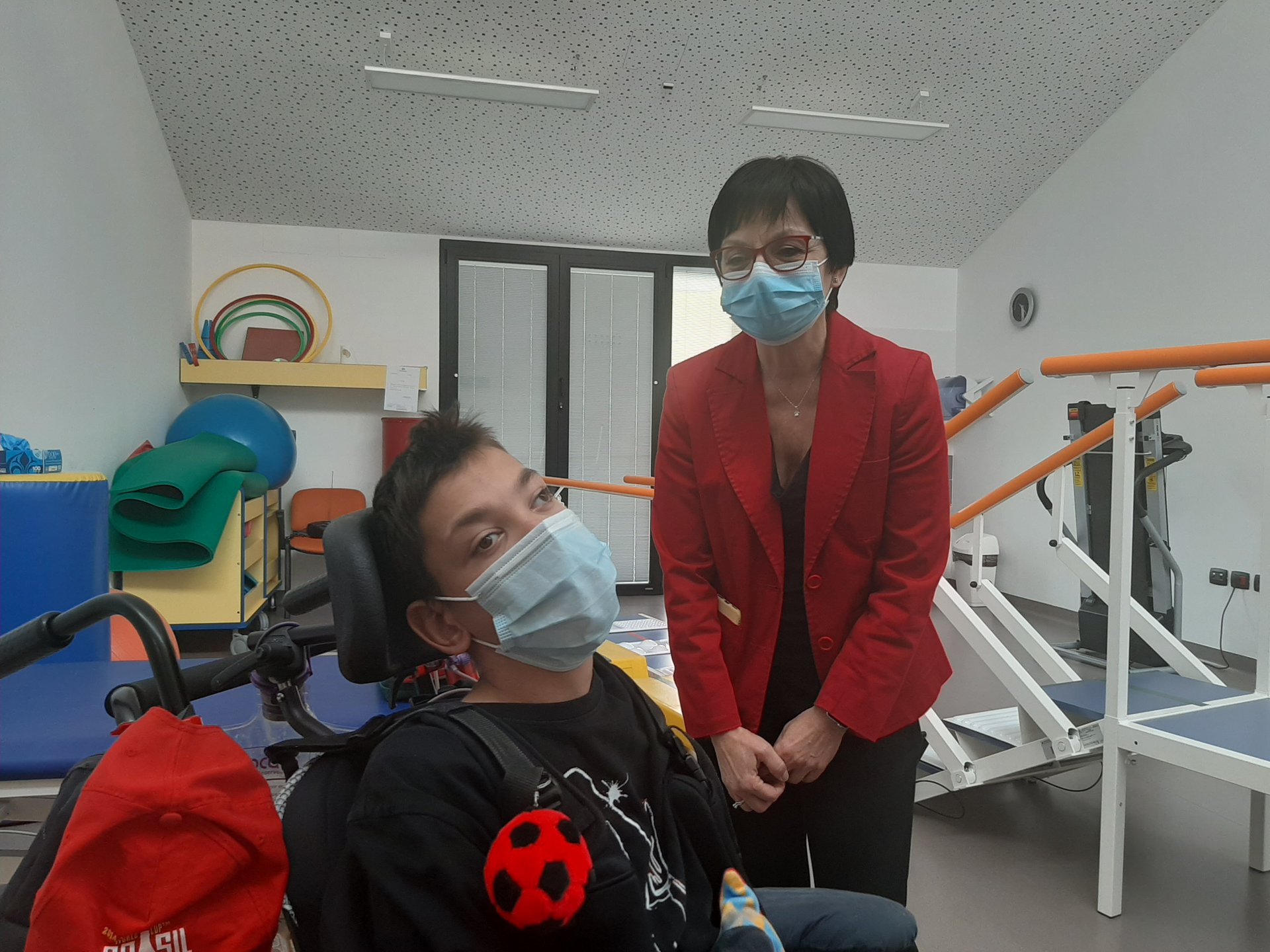 La Dottoressa Vigo è in piedi, sorridente con la mascherina. Davanti a lei un utente della Lega del Filo d'Oro, un ragazzo, anche lui con mascherina. I due si trovano all'interno di una palestra per la riabilitazione e dietro di loro ci sono gli strumenti per la riabilitazione.