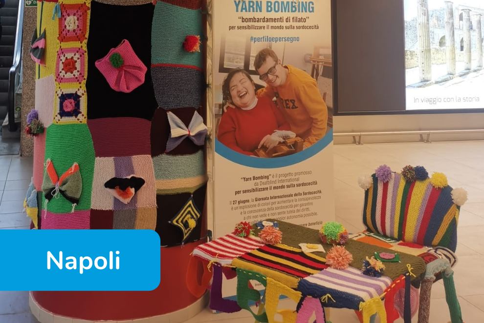 Installazione di “Yarn Bombing” in una stazione a Napoli, con seduta e colonna rivestite di colorati lavori all’uncinetto; sullo sfondo un banner della Lega del Filo d’Oro per la sensibilizzazione sulla sordocecità. 