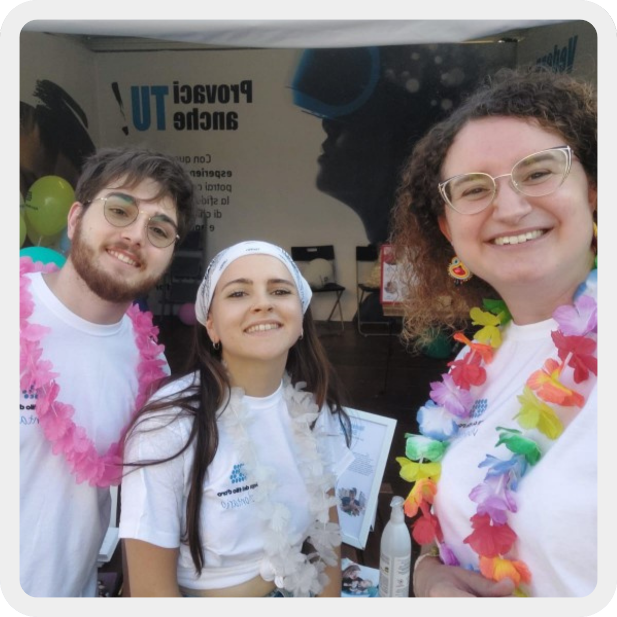 3 giovani volontari di Osimo sorridono di fronte allo stand di sensibilizzazione. Sono due ragazze ed un ragazzo. Indossano la t-shirt bianca con il logo Lega del Filo d'oro e delle collane hawaiiane. 