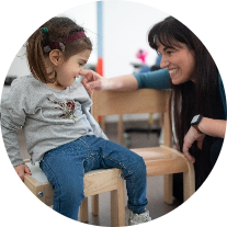 Mamma Alessandra e la piccola Letizia, bambina con la sindrome di Charge, giocano. Letizia seduta su una piccola sedia mentre la mamma è piegata sulle ginocchia e con ua mano le accarezza il viso.