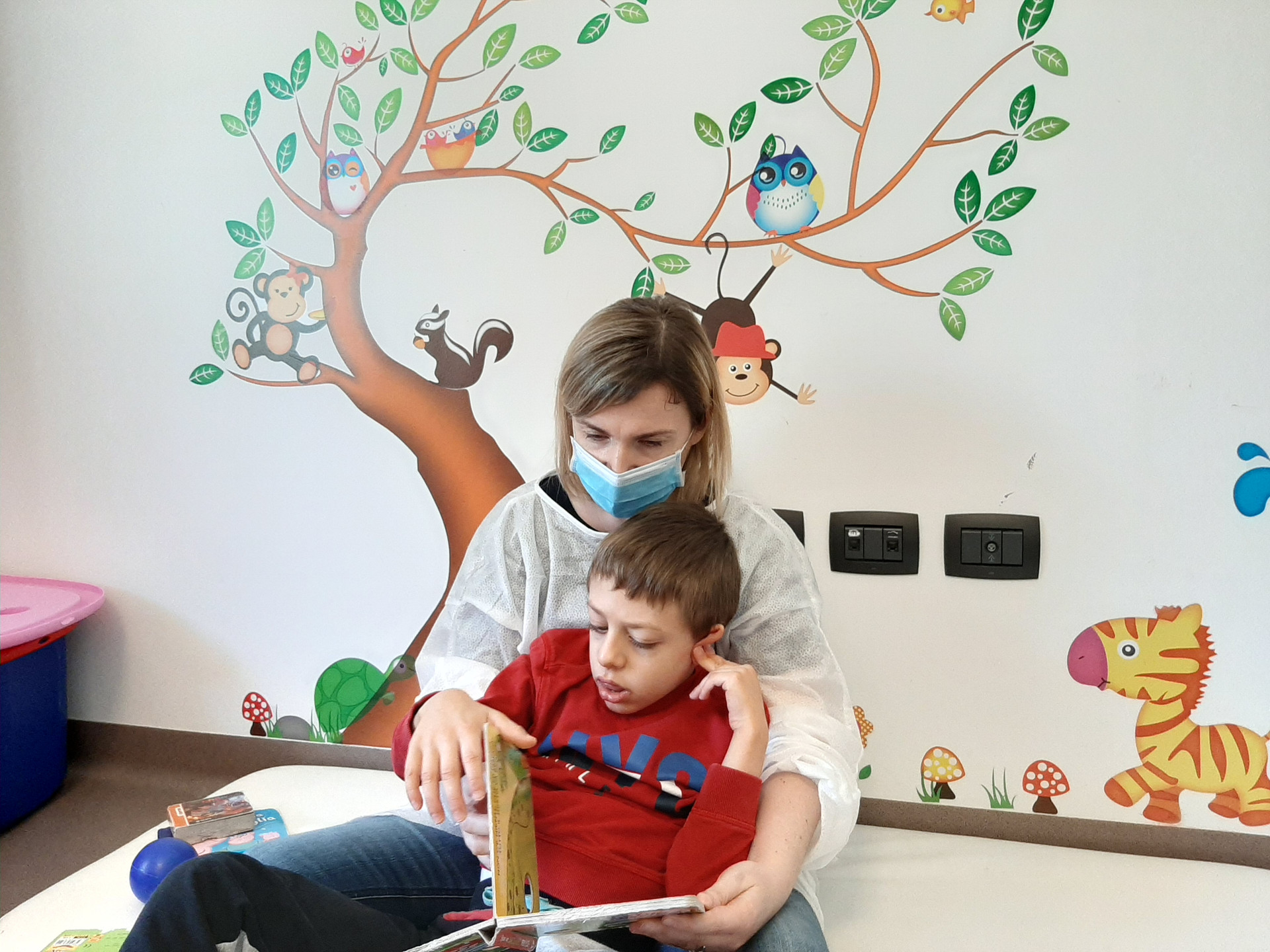 Mattia, piccolo utente del Centro di Osimo, siede per terra in braccio alla sua educatrice, che gli mostra un libro illustrato leggendogli una storia. L'educatrice indossa camice e mascherina.