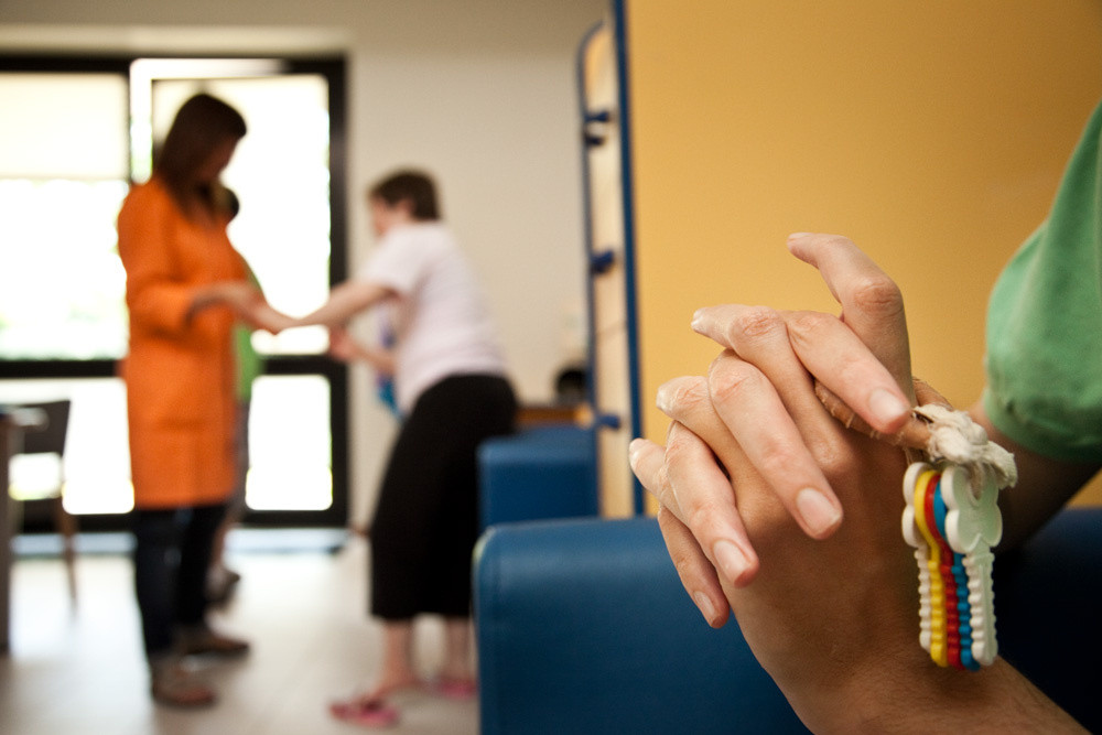 Primo piano di mani che reggono un mazzo di chiavi di plastica colorata. Sullo sfondo, sfocate, si intravedono due persone in piedi: un operatore che assiste un utente della Lega del Filo d'Oro.