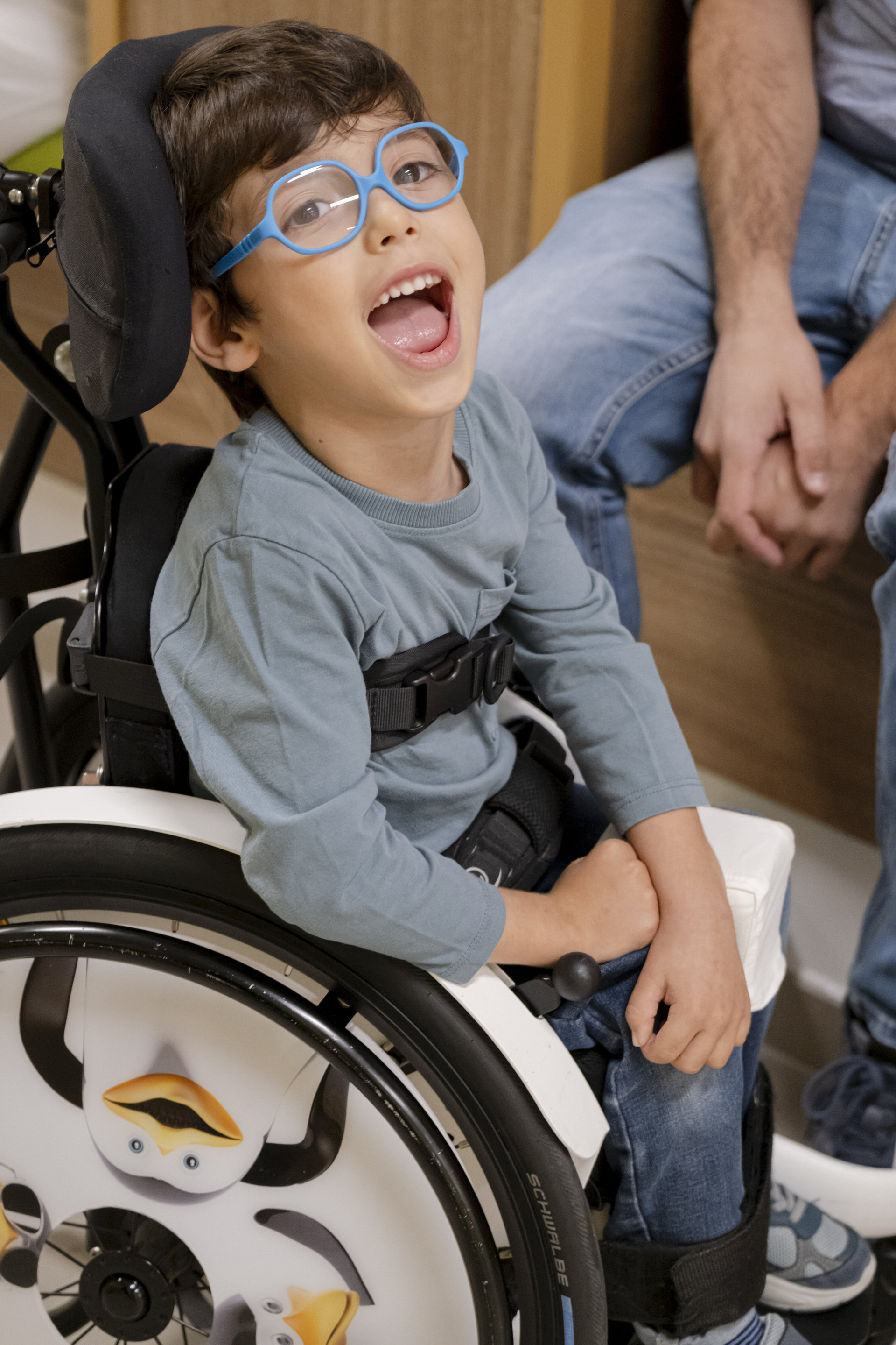 Nicola, seduto sulla sua sedie a rotelle con dei bellissimi Pinguini disegnati sulle ruote, ride mentre gli fanno una foto.