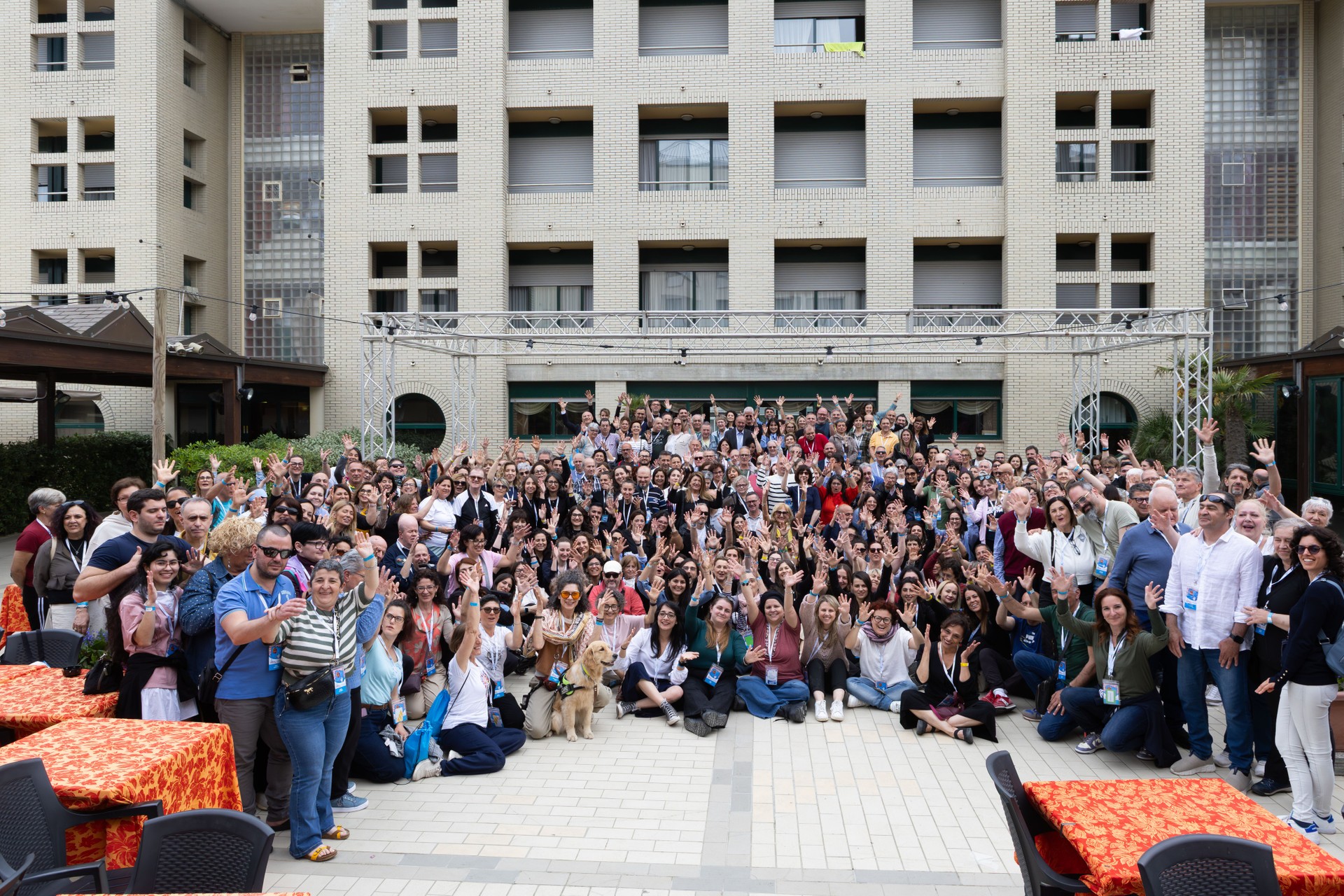 Questa foto racchiude tutti i partecipanti alla Conferenza a Cervia: persone con sordocecità, volontari, interpreti LIS, personale LFO, riunita nel piazzale esterno dell'Hotel Dante, luogo in cui si è tenuta la Conferenza. Ritrae persone sorridenti e con le braccia alzate in segno di festa, come se stessero applaudendo.