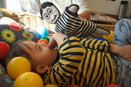 Edoardo è un bambino di circa quattro anni. Indossa una maglietta a righe gialle e nere ed è disteso dentro una cesta con cuscini a fiori, palline colorate (gialle, rosse, arancioni, blu verdi) e un pupazzo a righe bianche e nere che viene retto da un'operatrice di cui in foto c'è solo parte del braccio. 