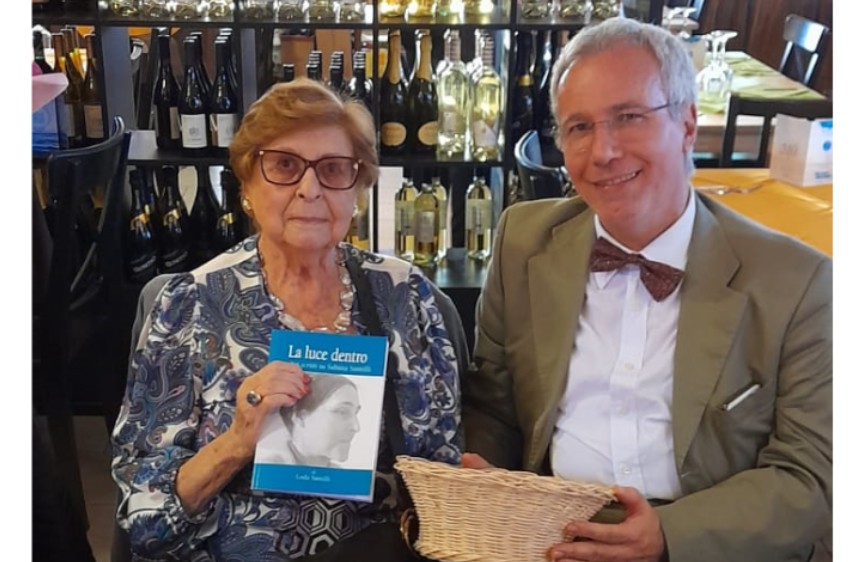 L'immagine mostra due adulti, una donna anziana e un uomo di mezza età, seduti l'uno accanto all'altra in un ambiente che sembra un negozio di vini, con numerose bottiglie sugli scaffali sullo sfondo. La donna tiene in mano un libro con il titolo "La luce dentro", mentre l'uomo, che indossa un papillon, tiene un piccolo cestino di vimini e sorride.