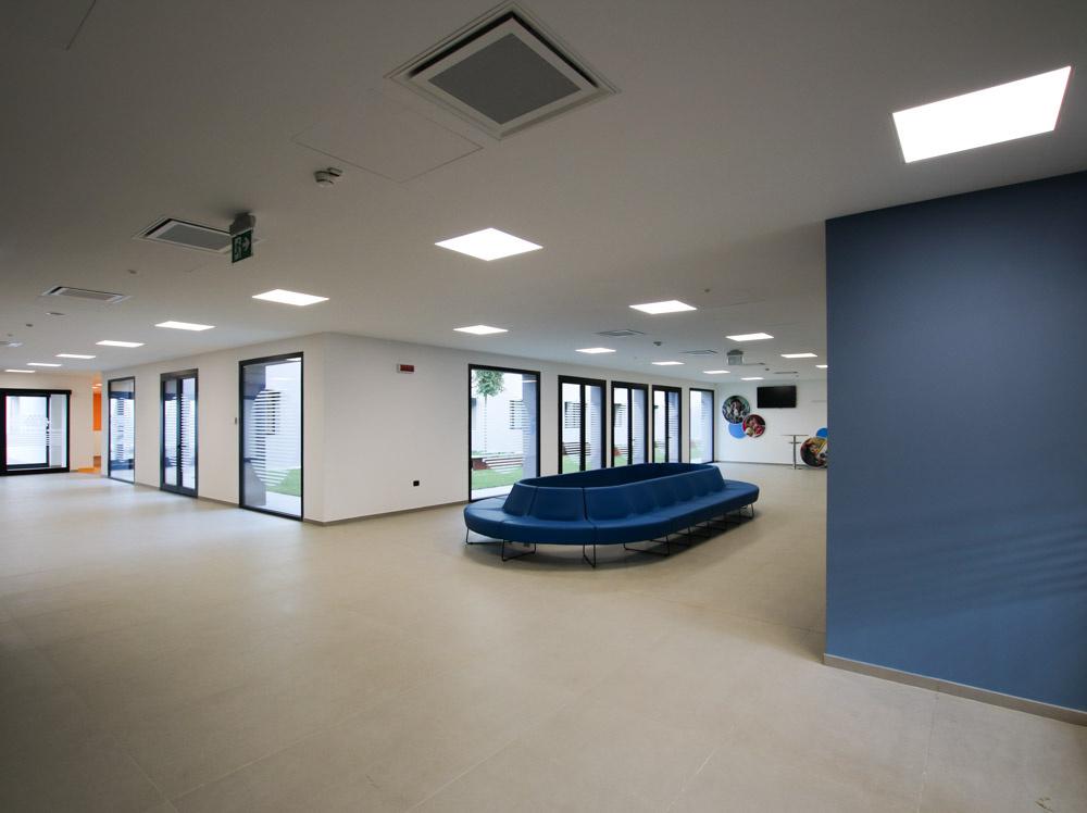 L'atrio d'ingresso del primo lotto del Centro Nazionale. Un ambiente ampio e con illuminazione diffusa, al centro un divano ovale blu con molti posti a sedere per l'accoglienza di ospiti e famiglie in arrivo. ampie vetrate si aprono su uno dei giardini interni del Centro