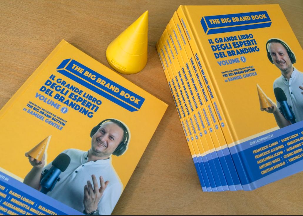 Pile del libro “The Big Brand Book – Il grande libro degli esperti del branding” con copertina gialla e foto di Samuel Gentile con microfono; in primo piano anche un cono giallo simbolico. 