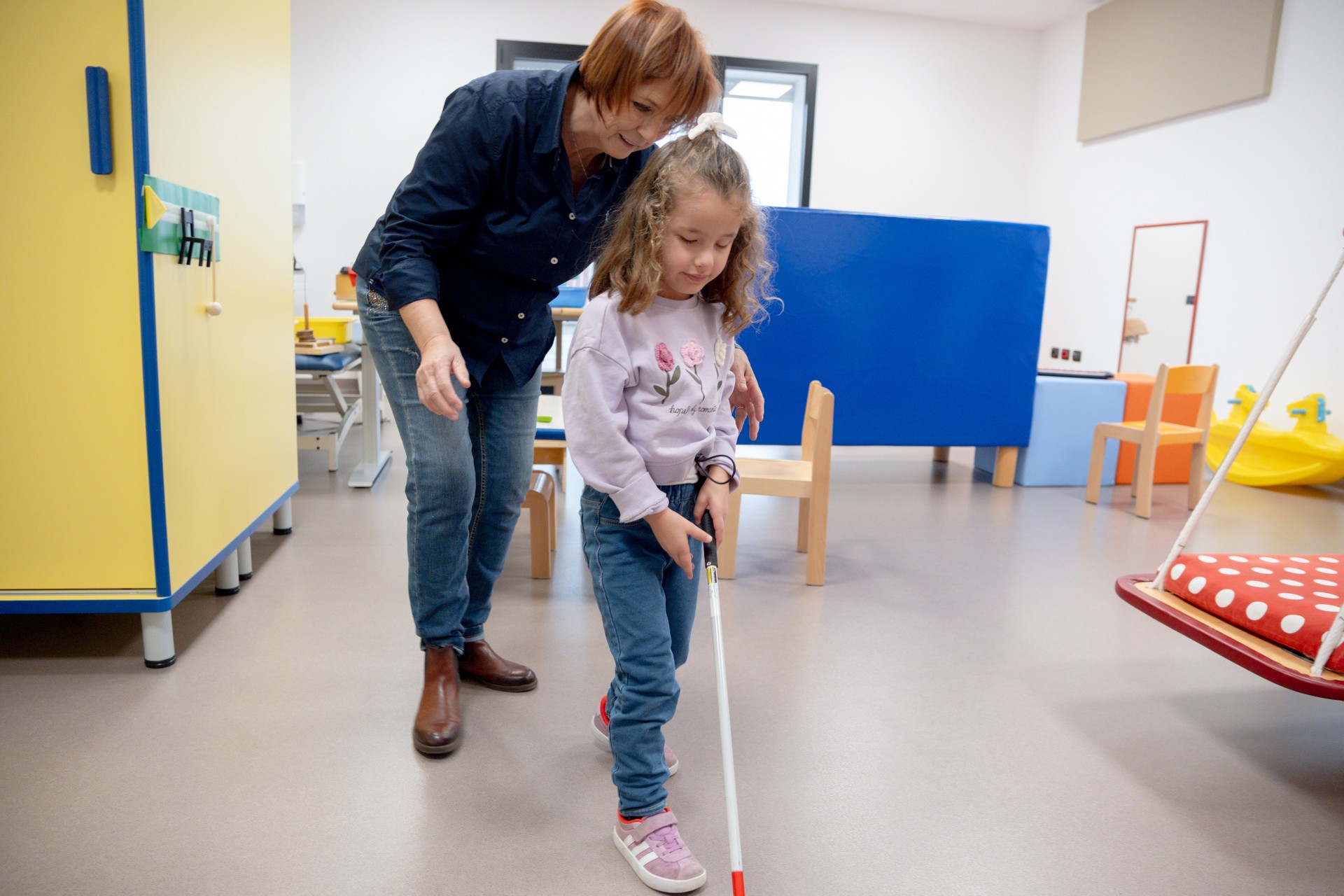 Chiara è insieme alla sua educatrice, che la aiuta a camminare. Chiara infatti sta camminando utilizzando il bastone bianco e rosso per evitare gli ostacoli davanti a lei