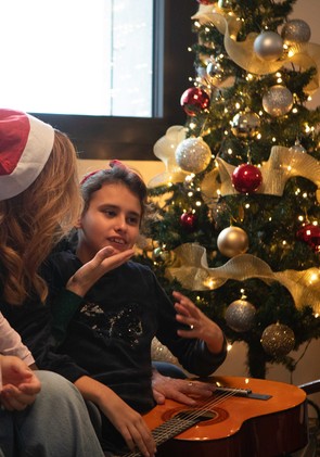 Aurora, bambina con pluridisabilità, riceve una carezza dalla sua educatrice. Sono sedute vicino all'albero di Natale e tengono sulle gnocchia una chitarra in modo che Aurora possa sentire le vibrazioni della musica