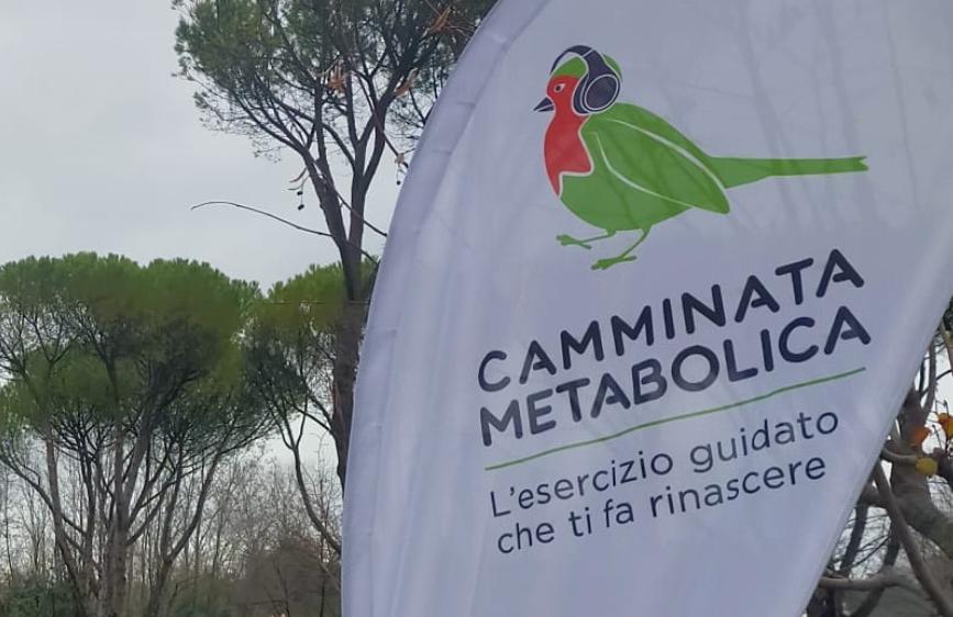 L'immagine mostra una parte di un banner bianco che promuove una "CAMMINATA METABOLICA", visibile all'aperto con degli alberi sullo sfondo. Il banner presenta il logo di un uccellino verde con il petto rosso e delle cuffie, e sotto il testo "L'esercizio guidato che ti fa rinascere".