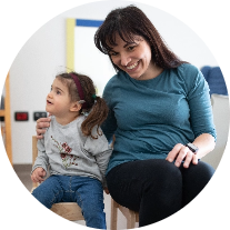 Mamma Alessandra e la piccola Letizia, bambina con la sindrome di Charge, sono sedute su delle piccole sedie e sorridono.