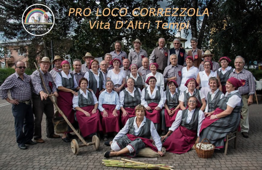 L'immagine mostra un numeroso gruppo di persone, il "Gruppo Antichi Mestieri Pro Loco Correzzola", posato all'aperto, con un logo in alto a sinistra. Molti indossano abiti tradizionali o costumi che richiamano epoche passate, con gonne lunghe rosse, grembiuli e copricapi, e alcuni tengono attrezzi agricoli o cesti, evocando un'atmosfera di "Vita D'Altri Tempi".