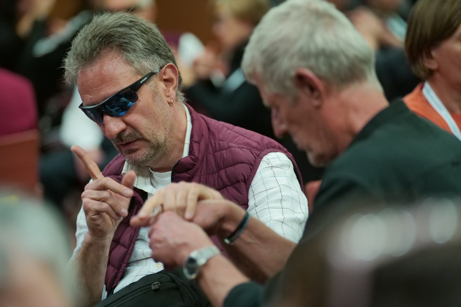 Due utenti della Lega del Filo d’Oro sono ritratti in un momento informale. Uno, in primo piano, indossa occhiali sportivi, camicia a scacchi bianca e gilet bordeaux. L’altro, leggermente sfocato e di spalle, indossa una camicia verde.