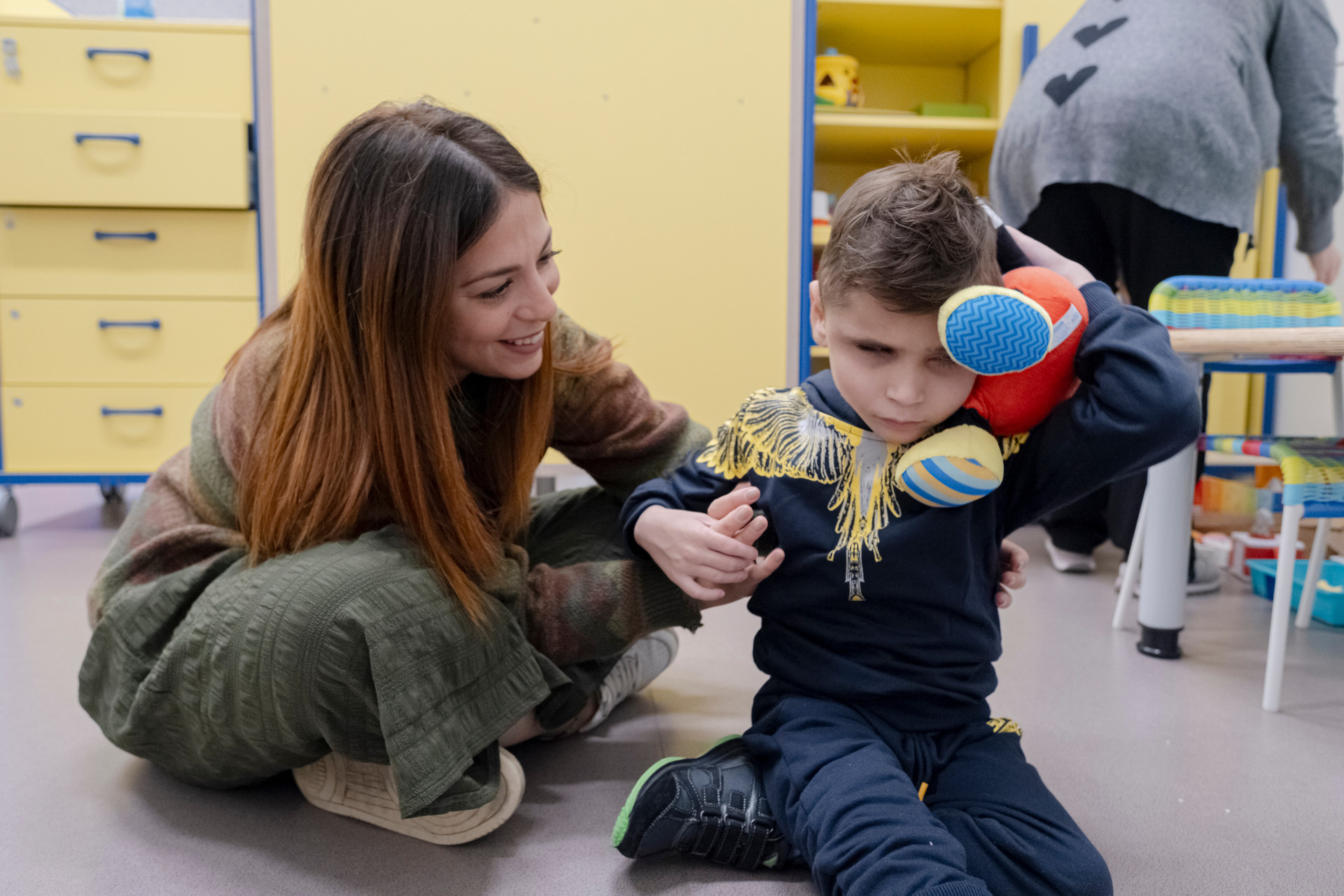 Foro di Matteo, un bambino sordocieco insieme all'assistente sociale della Sede Territoriale di Napoli. Entrambi sono seduti per terra. Matteo tiene in mano un peluche colorato, l'assistente sociale sorride