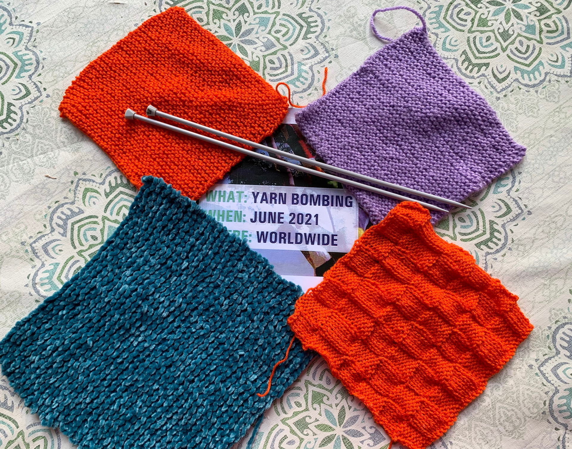 Quattro pezze quadrate colorate sono appoggiate su un piano ricoperto con una tovaglia bianca con decorazioni verdi. Le pezze sono disposte in cerchio (con i vertici che si toccano, essendo quattro quadrati) e sono di colore rosso, viola, rosso e blu. Nello spazio centrale lascio dalla disposzione delle pezze c'è un biglietto che recita "WHAT: YARN BOMBING WHEN: JUNE 2021 WHERE: WORLDWIDE". Su due pezzoline sono appoggiati due ferri da uncinetto.