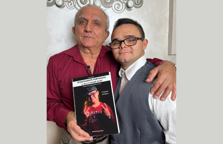 Due uomini sono abbracciati e sorridono davanti alla fotocamera; l’uomo a sinistra, Arnaldo, tiene in mano un libro con la copertina nera. A destra Marco Baruffaldi, vestito con camicia bianca e gilet grigio. Sul libro c’è la foto di Marco con il titolo “Sfigatamente Fortunato – Bello essere fuori”.