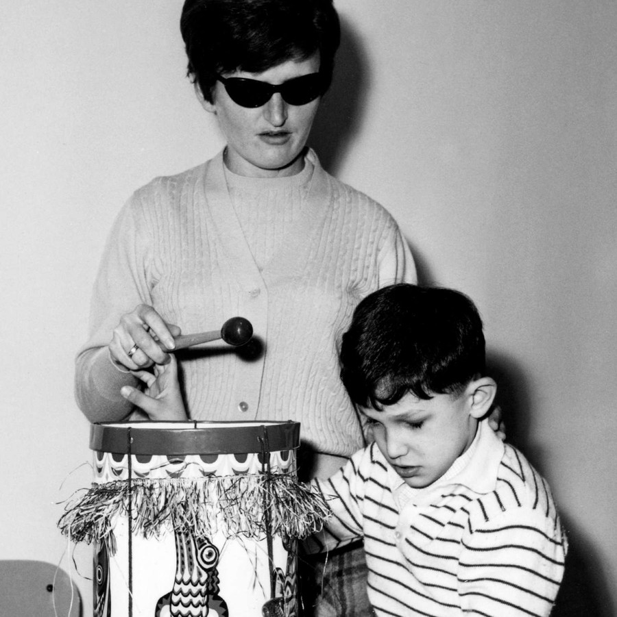 Foto in bianco nero, scattata a fine anni '60. Un bambino sordocieco gioca con un tamburo, aiutato da un'operatrice.