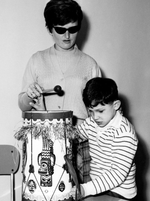 Foto in bianco nero, scattata a fine anni '60. Un bambino sordocieco gioca con un tamburo, aiutato da un'operatrice.