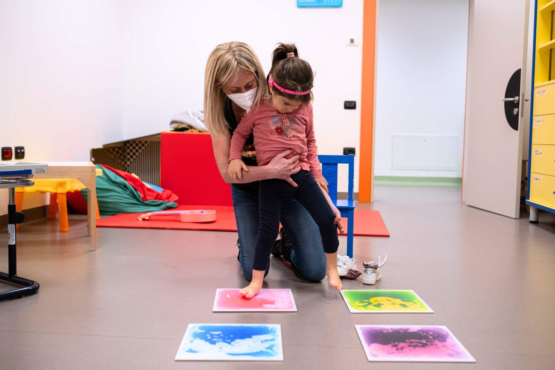 Letizia, una bambina di circa 10 anni con la sindrome di Charge, fa attività con l'educatrice: con i piedi nudi cammina sopra delle mattonelle colorate per sentire a contatto con la pelle la texture di queste.