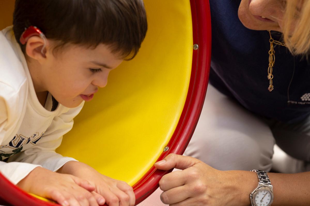 Agostino, bambino con sindrome di Charge seguito al Centro di Osimo, sta sdraiato dentro un grande tubo giallo e rosso, al suo fianco la fisioterapista Francesca