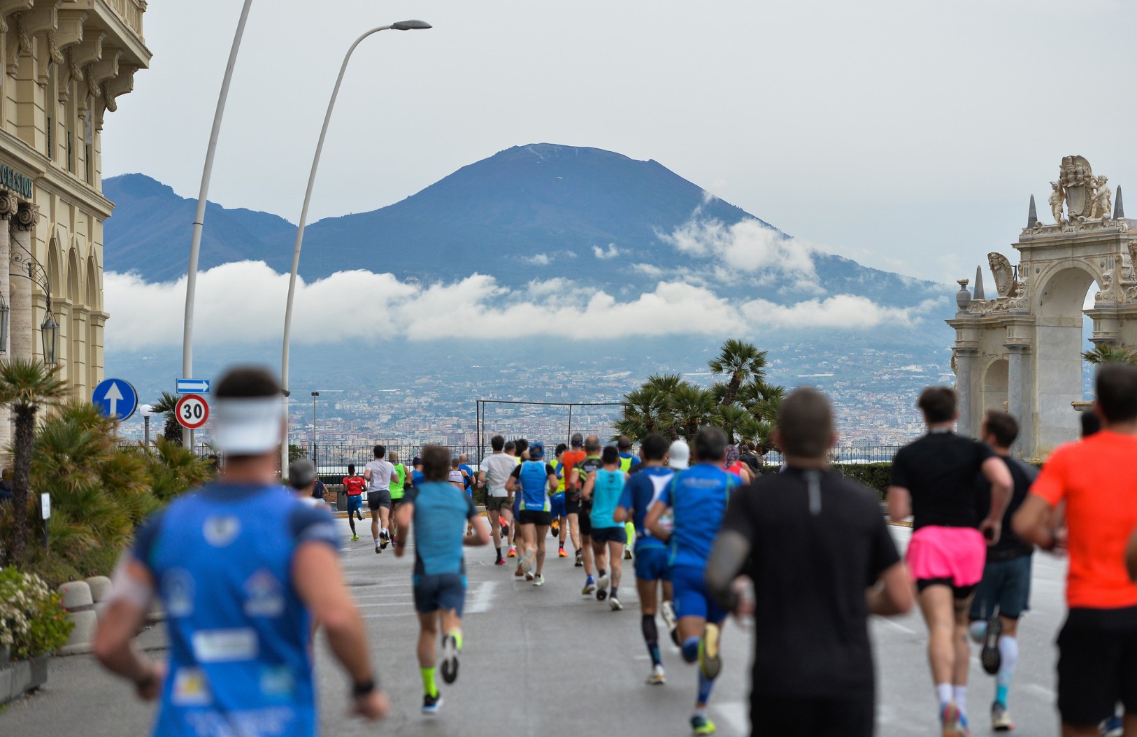 Foto di persone che corrono durante la maratona a Napoli. Sullo sfondo si vede il Vesuvio