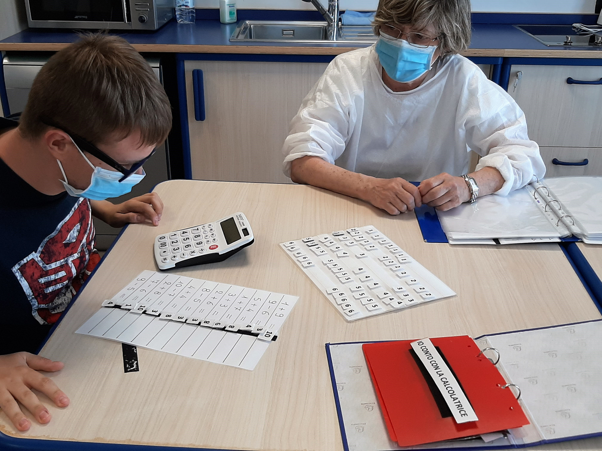 Victor durante una lezione di matematica con la sua educatrice. Usa una calcolatrice per compiere le operazioni indicate dall'educatrice su un foglio, e con un velcro attacca il risultato scritto su un foglietto. Victor e l'educatrice indossano mascherine chirurgiche.