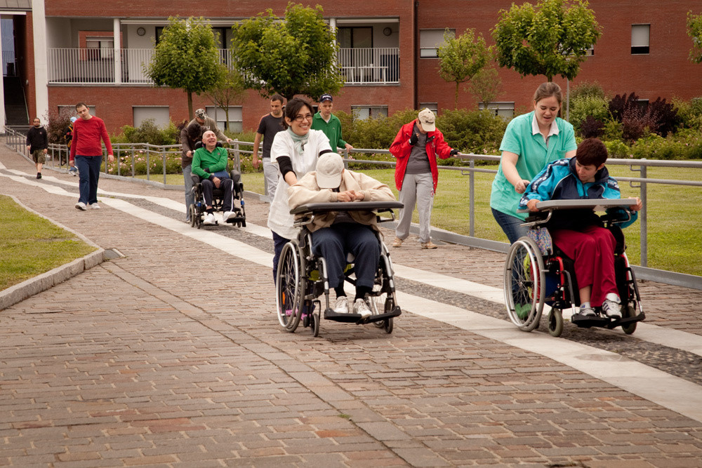 Alcuni utenti passeggiano per i percorsi esterni di un Centro Residenziale. 3 con disabilità motorie sono spinti dalle operatrici.