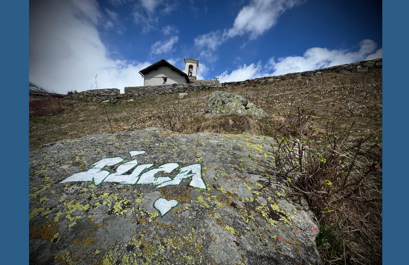 Scorcio suggestivo di una piccola chiesa di montagna su una collina erbosa sotto un cielo nuvoloso. In primo piano, su un grande masso ricoperto di muschio, si nota la scritta bianca "LUCA" seguita da un cuore, dipinta in stile graffiti. Sullo sfondo, un muretto a secco delimita l’edificio. 