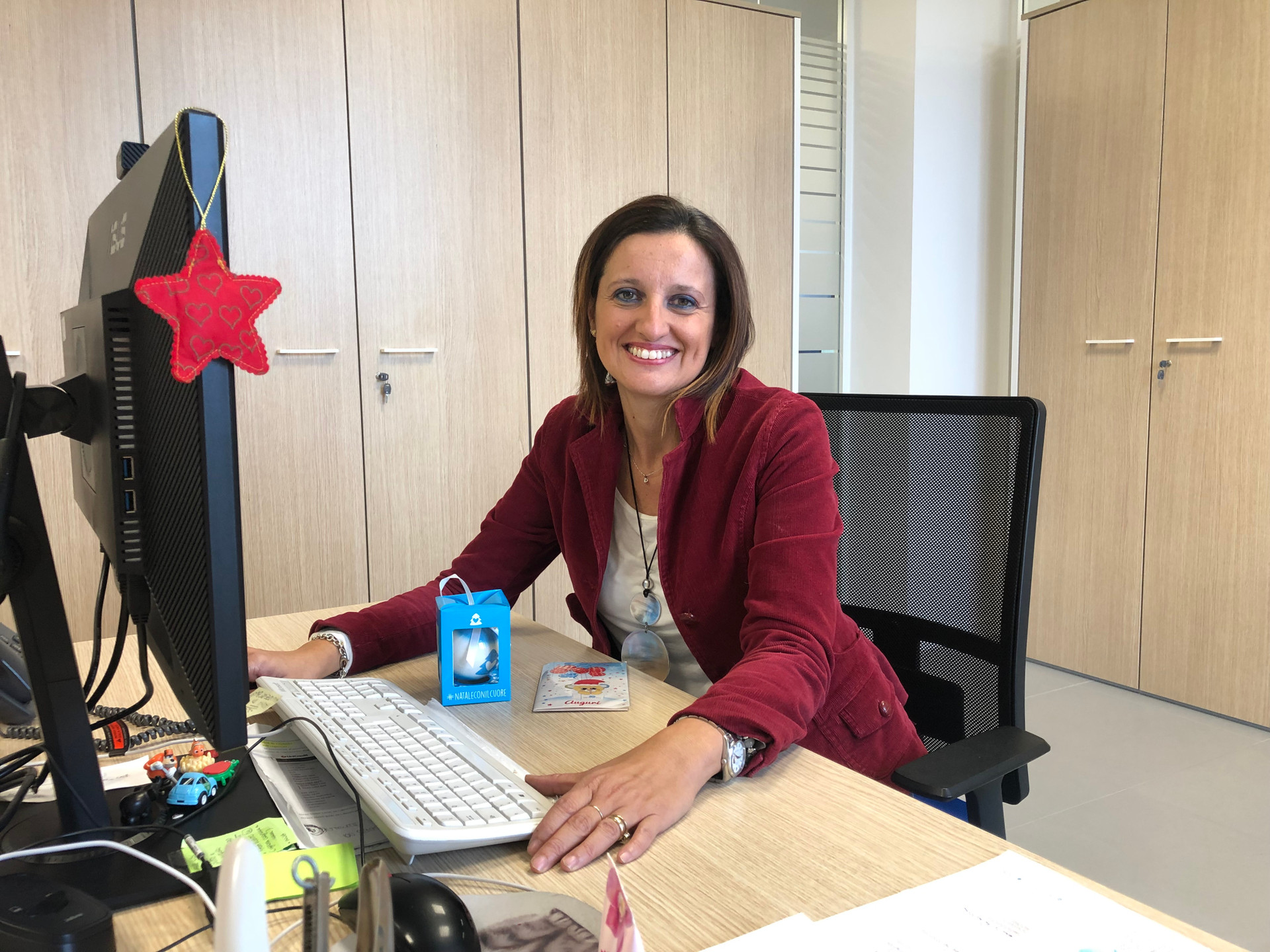Francesca è seduta alla sua scrivania, indossa una giacca rosso scuro ed è sorridente.