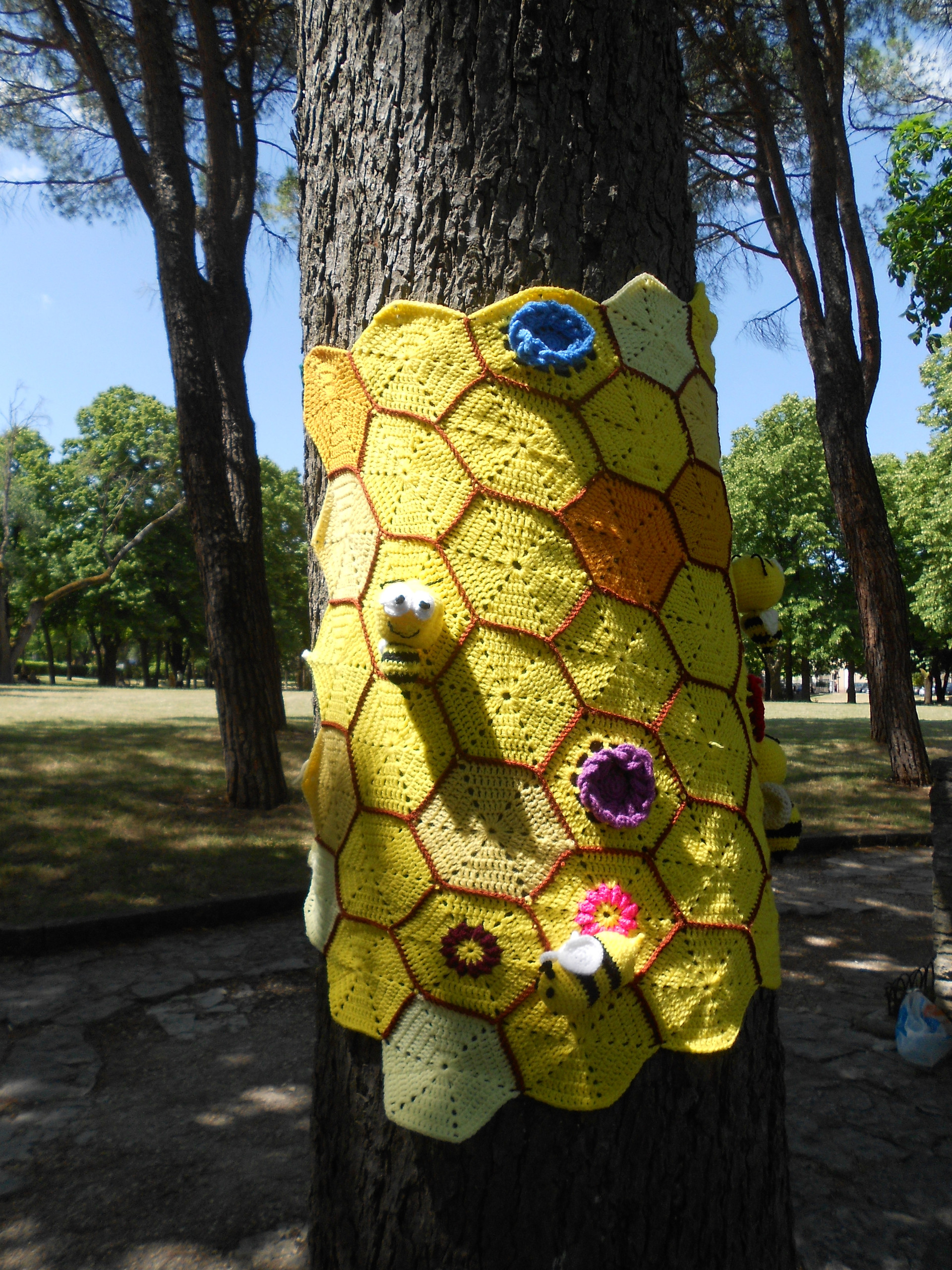 Foto di un lavoro di Yarn Bombing all'uncinetto che abbraccia un albero, creando un effetto colorato e artistico.