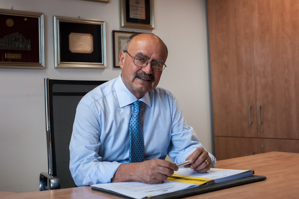 Il Presidente Rossano Bartoli seduto alla sua scrivania nel suo ufficio. Davanti a lui è aperto un grosso quaderno e lui tiene tra le mani una penna.