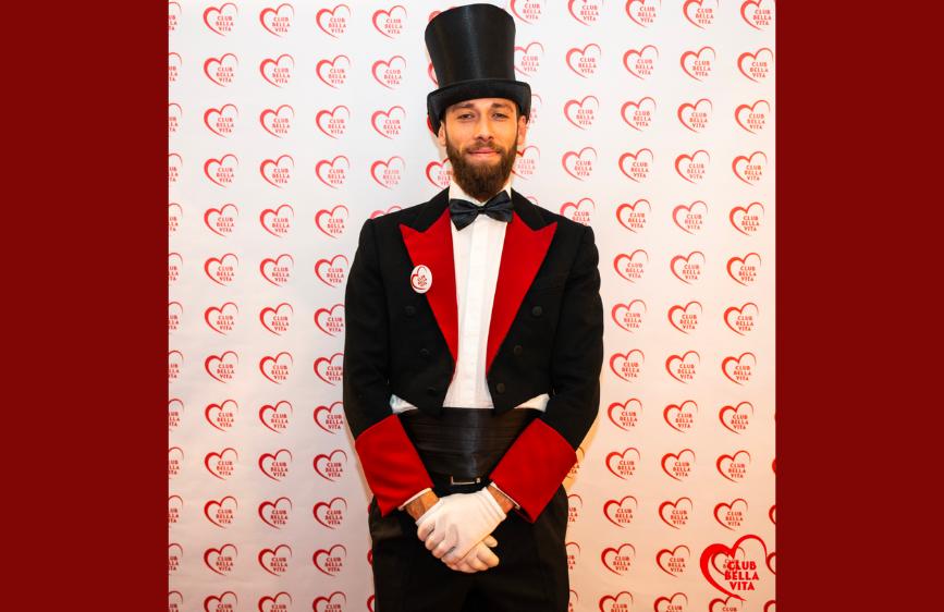 Nella foto è ritratto un uomo in posa frontale, vestito con un elegante frac nero con ampi risvolti rossi, guanti bianchi, papillon e un cilindro nero in testa. Indossa un distintivo con la scritta "Club della Vita" su sfondo bianco e rosso. Alle sue spalle, il fondale è completamente ricoperto da un motivo ripetuto di cuori rossi con la stessa scritta. L'inquadratura è centrata e l’illuminazione uniforme, adatta a un contesto formale o celebrativo.